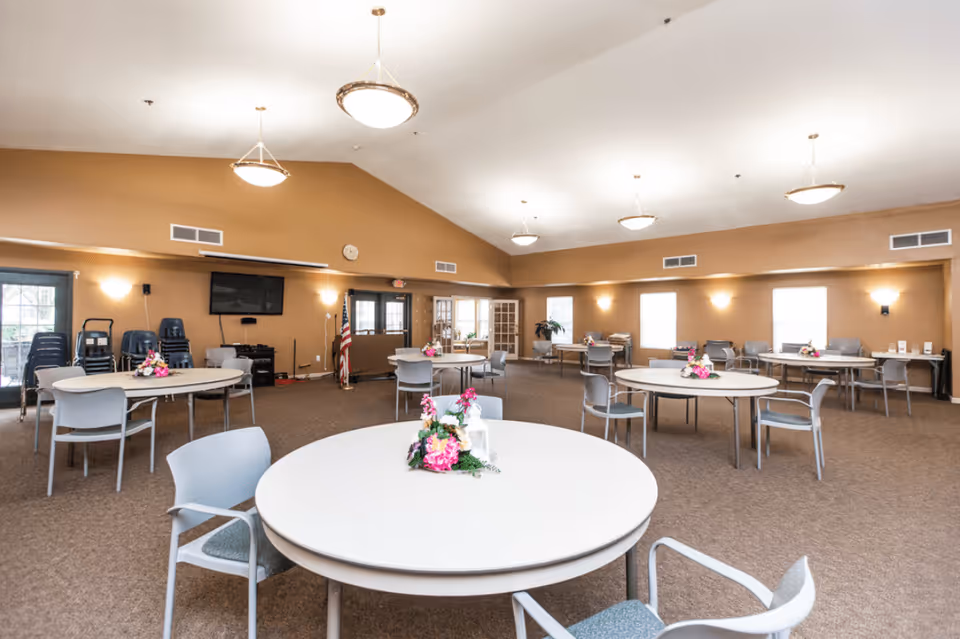 Spacious multipurpose dining/activity room with round tables and chairs, ceiling lights, and floral centerpieces.