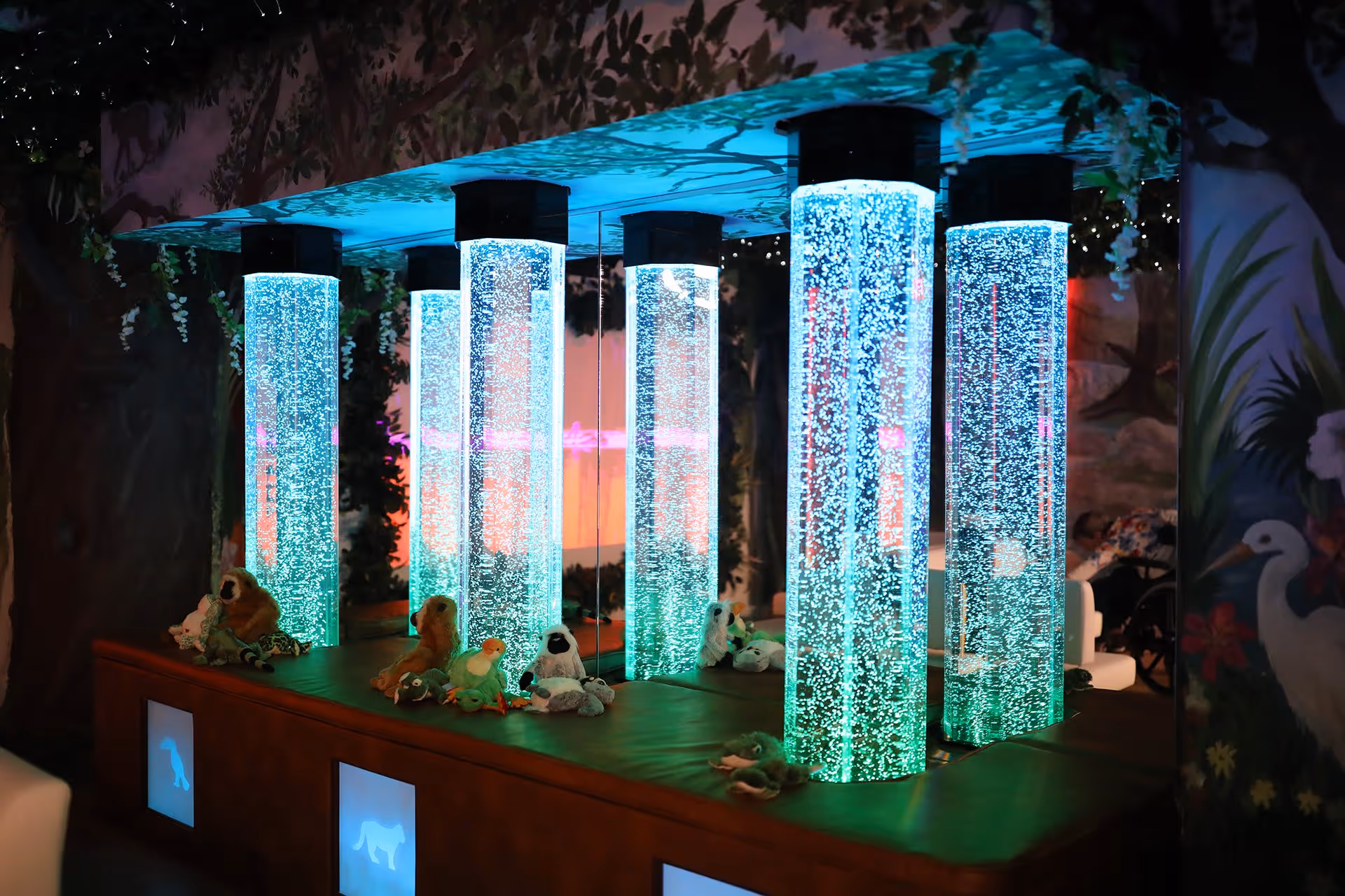 A sensory room featuring six illuminated bubble tubes emitting blue and green light, surrounded by plush animal toys on a padded bench. The background includes nature-themed wall murals with trees and birds, creating a calming environment.