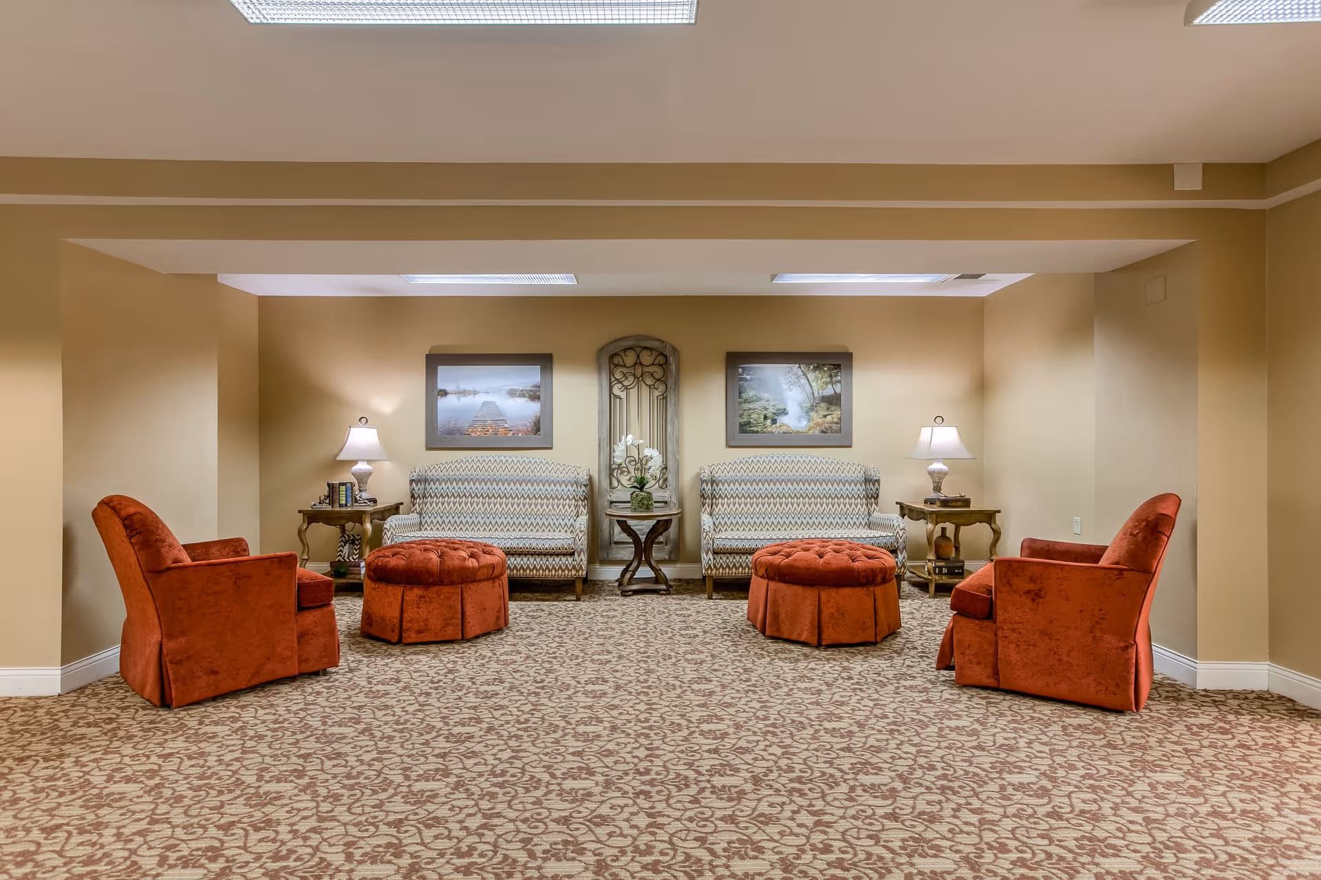 A cozy sitting area in a retirement community with two patterned sofas, two red armchairs, and two round red ottomans arranged on a patterned carpet. There are two side tables with lamps and books, and two framed landscape pictures on the beige walls.