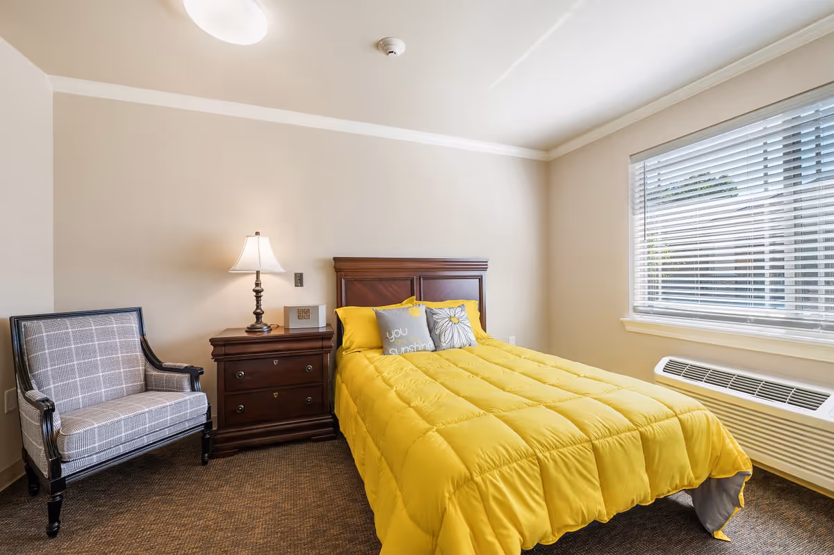 A bedroom with a bed covered in a bright yellow comforter and two decorative pillows, one with the text 'you sunshine'. Next to the bed is a wooden nightstand with a lamp and a small decorative box. To the left of the nightstand is a cushioned armchair with a plaid pattern. A large window with white blinds is on the right wall, and a heating/cooling unit is beneath the window.