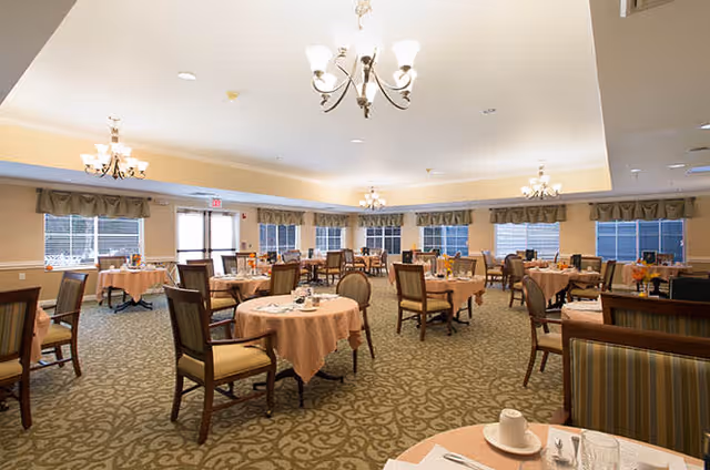 A spacious dining room with round tables covered in peach-colored tablecloths, each set with cups, saucers, and silverware. The room features multiple chandeliers hanging from the ceiling, large windows with valances, and patterned carpet flooring. Chairs with cushioned seats and backs are arranged around the tables.