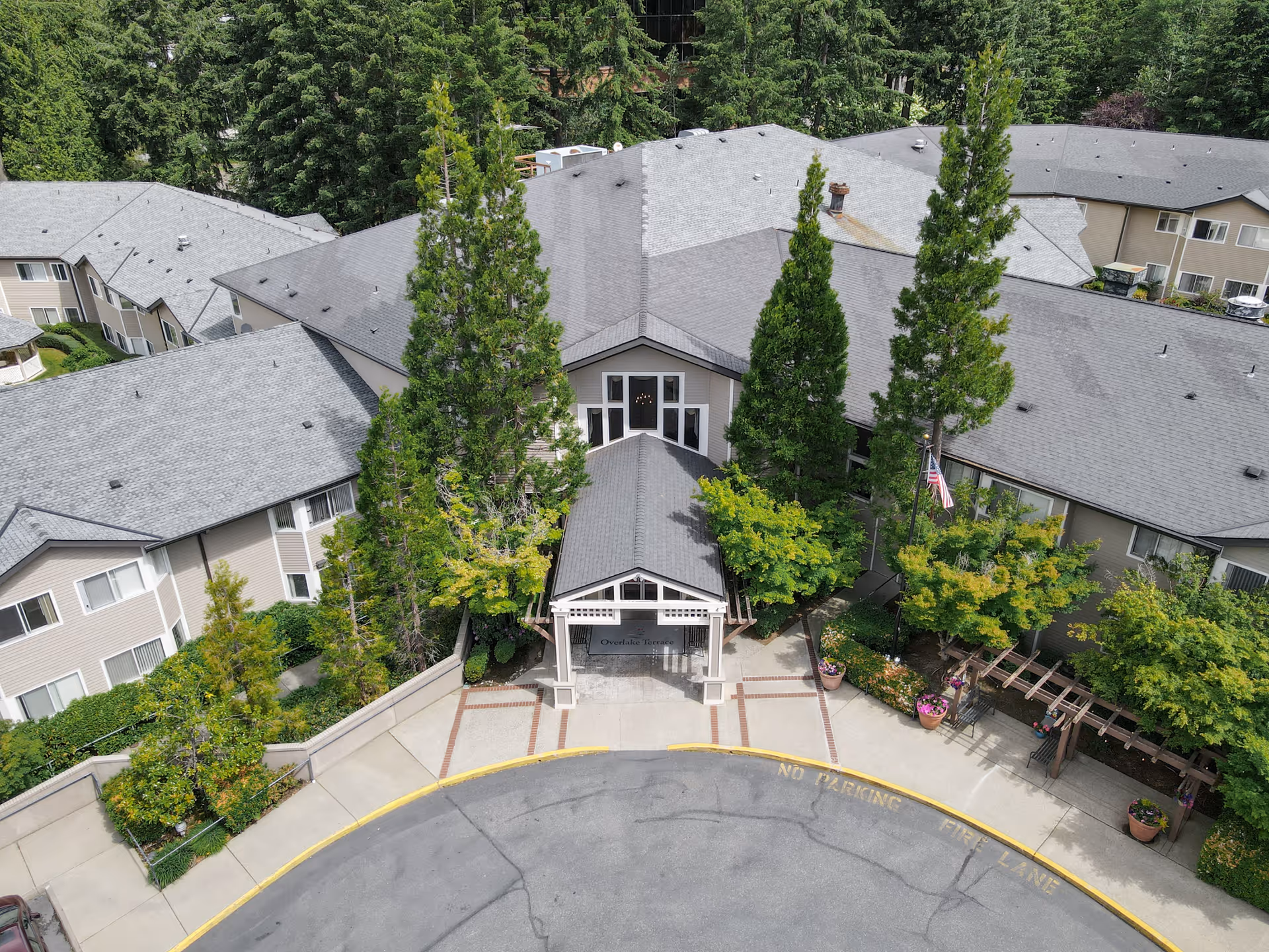 Aerial view of Overlake Terrace Assisted & Senior Living facility showing the main entrance with a covered walkway, surrounded by tall trees and landscaped greenery. The building has a gray roof and beige exterior walls, with a circular driveway marked with 'No Parking Fire Lane'.