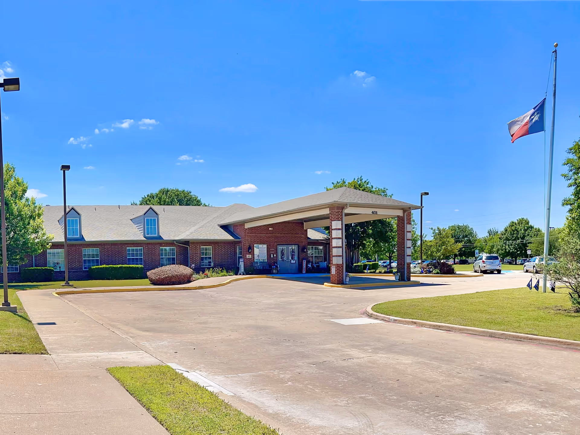 Front entrance of a single-story brick senior living building with a covered drop-off porte-cochère, driveway, parking and a Texas flag flying.