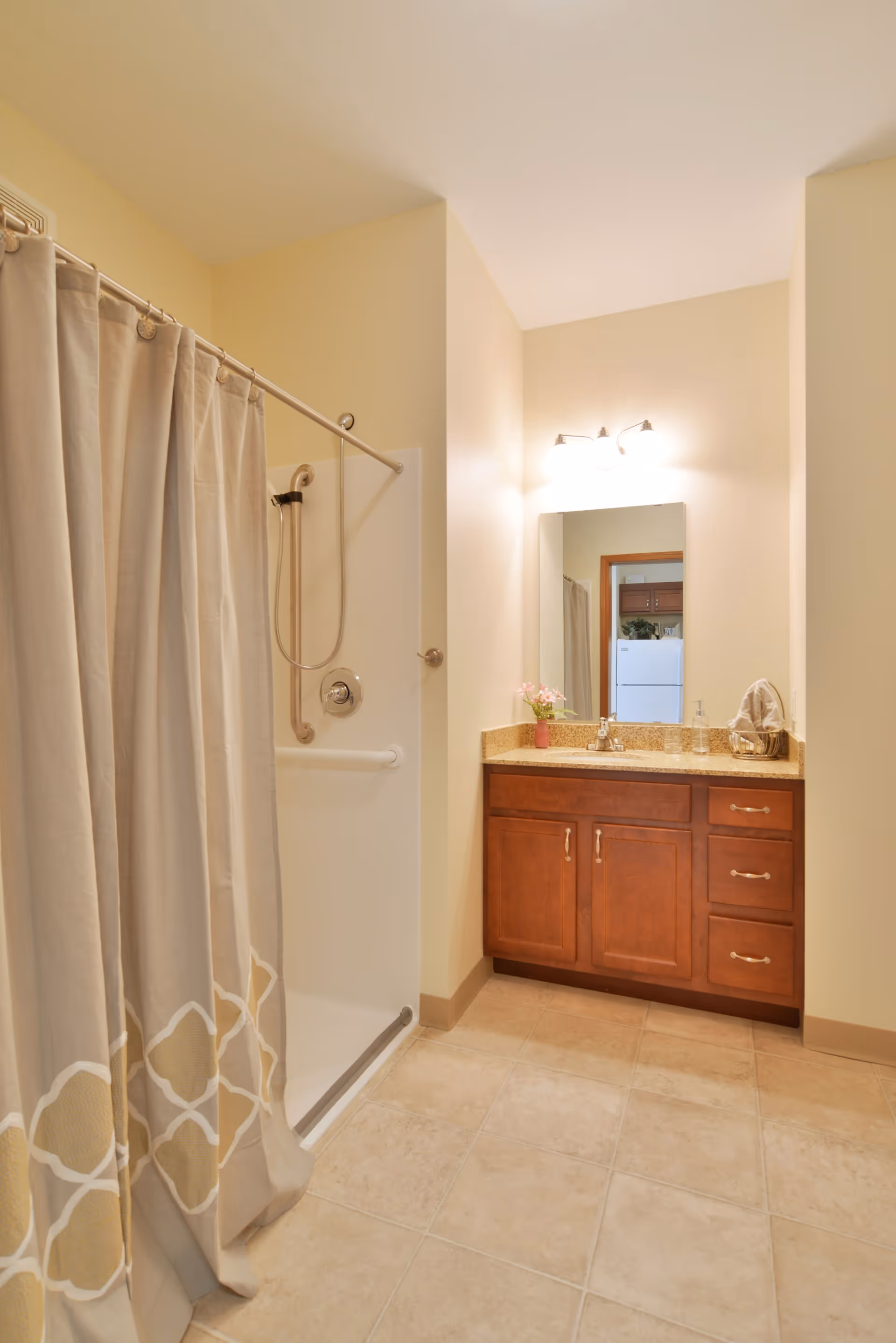 A clean and well-lit bathroom featuring a shower with a beige patterned curtain on the left and a wooden vanity with a granite countertop and a mirror above it on the right. The floor is tiled, and the walls are painted a light beige color.