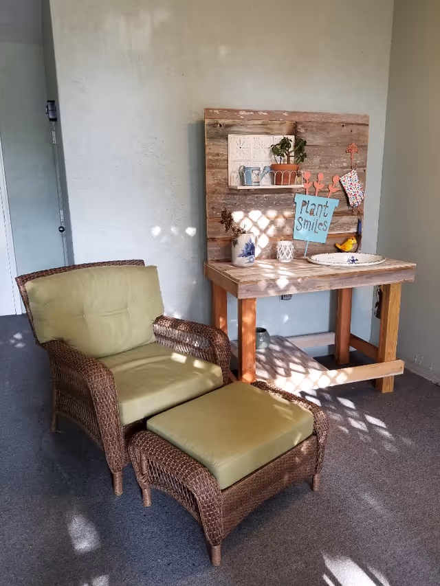 A cozy corner with a wicker armchair and matching ottoman, both with light green cushions, next to a rustic wooden table with decorative items including a small plant, watering cans, a ceramic pitcher, and a sign that reads 'Plant Smiles'. The room has light-colored walls and carpeted floor with sunlight casting shadows.