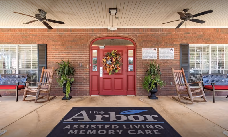 Front entrance of The Arbor Assisted Living and Memory Care facility featuring a red double door decorated with a fall-themed wreath, two large potted plants on either side, two ceiling fans above, and a black welcome mat with the facility's name. There are two wooden rocking chairs and two benches with red cushions on either side of the entrance.