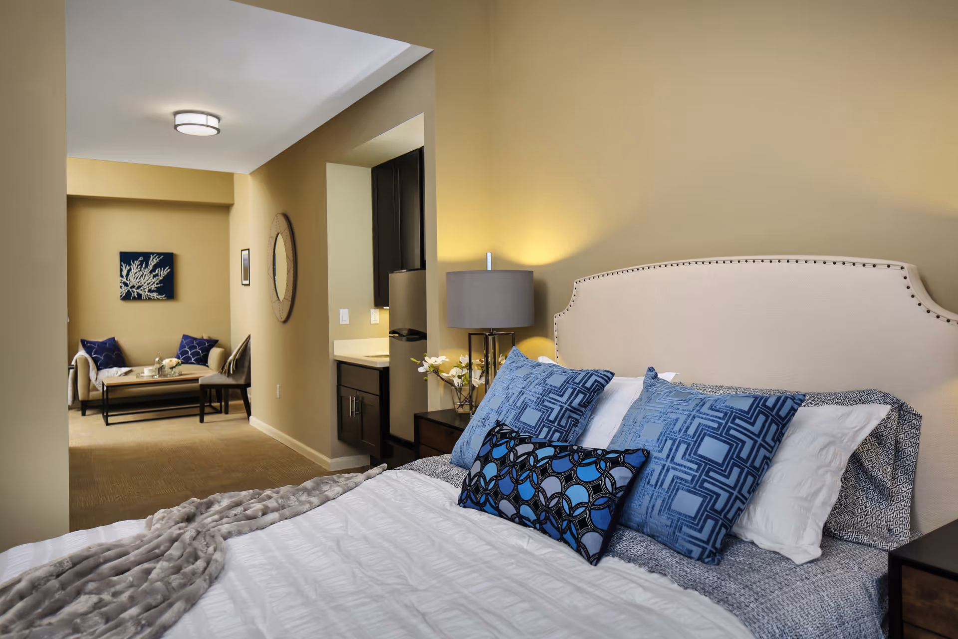 A furnished bedroom with a large bed topped with blue pillows and a bedside lamp, opening into a small sitting area with a sofa and coffee table.
