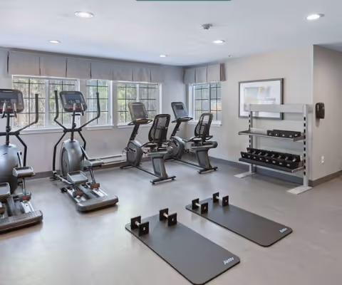 A fitness room with exercise equipment including two elliptical machines, two recumbent stationary bikes, two black exercise mats on the floor, and a rack holding various dumbbells. The room has large windows with gray valances, light gray walls, and a framed picture on one wall.
