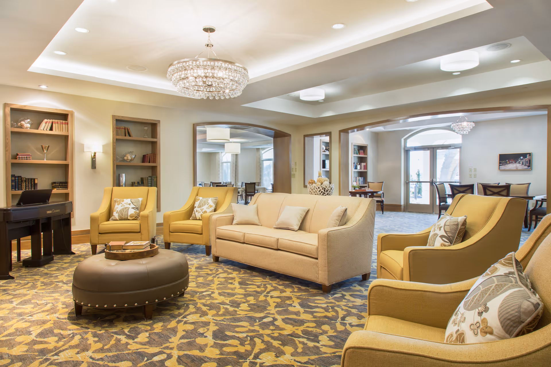 A bright and spacious living room area with beige and yellow upholstered chairs and a beige sofa arranged around a round brown ottoman. The room features built-in bookshelves with books and decorative items, a chandelier, and large windows letting in natural light. In the background, there is a dining area with tables and chairs visible through large open doorways.