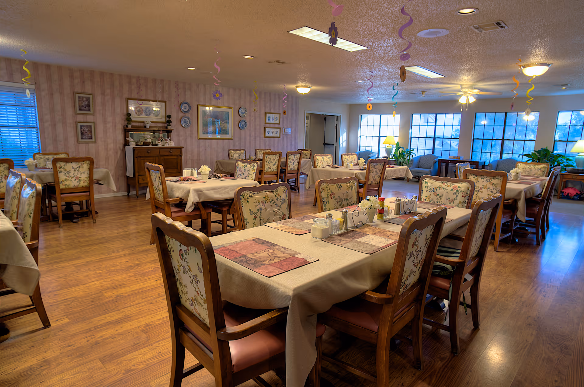 A spacious dining room in an assisted living facility with multiple tables covered in beige tablecloths and floral cushioned chairs. The room has wooden flooring, soft lighting from ceiling fixtures and lamps, and large windows letting in natural light. Decorative plates and framed pictures adorn the walls, and colorful hanging decorations are suspended from the ceiling.