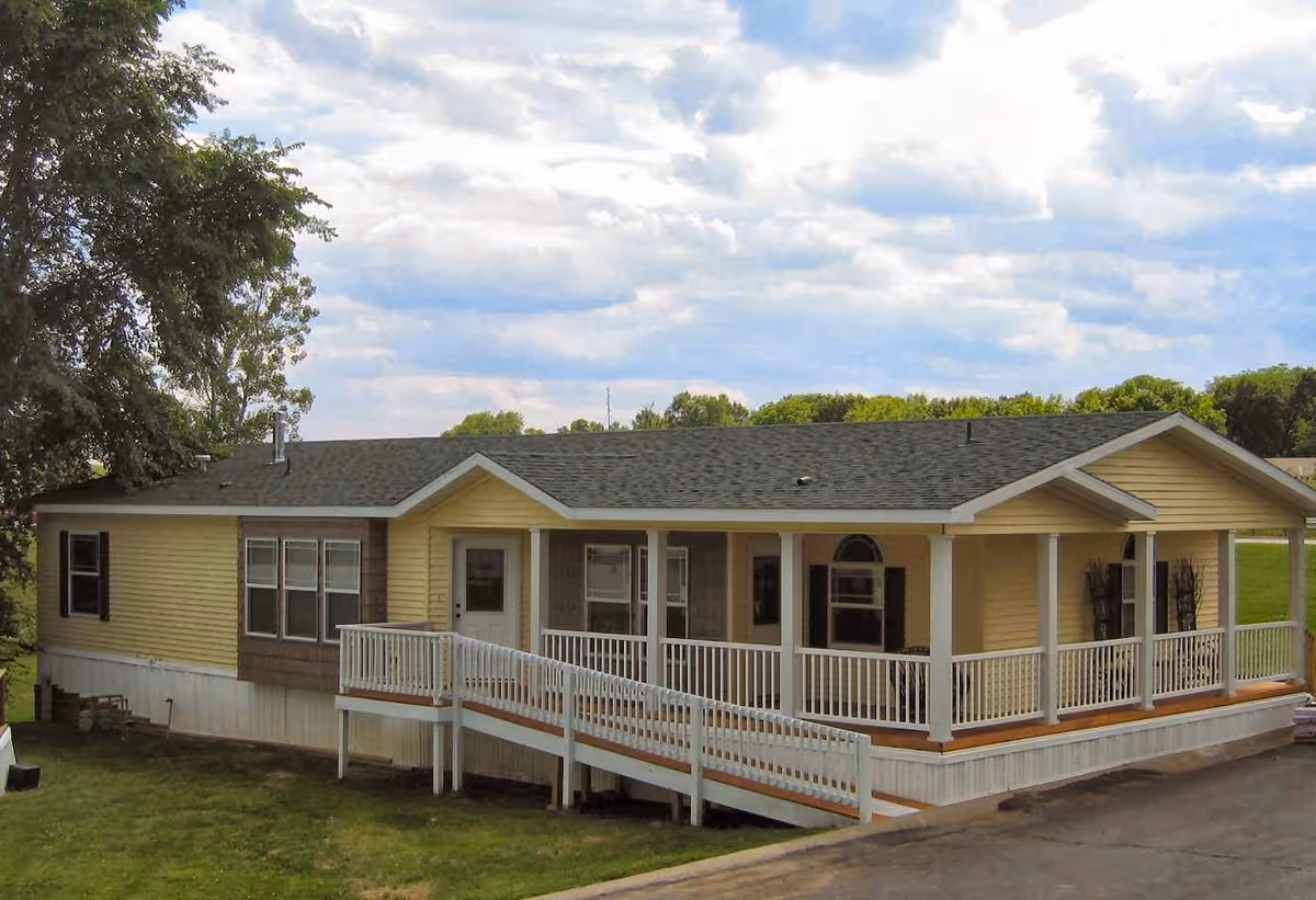 Single-story yellow manufactured home with a covered porch and wheelchair ramp on a grassy lot under a cloudy sky.