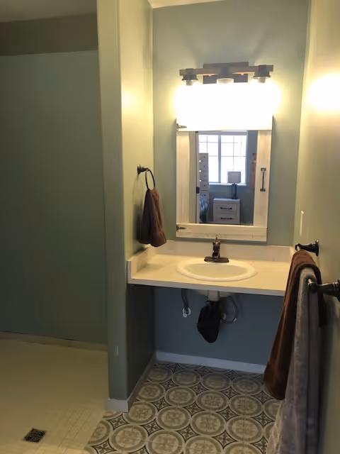 A bathroom sink area with a white countertop and an oval sink. Above the sink is a rectangular mirror with a white frame and a light fixture with four bulbs. There are brown towels hanging on towel rings and bars on the walls. The floor has patterned tiles, and to the left is a walk-in shower area with a tiled floor and a drain.