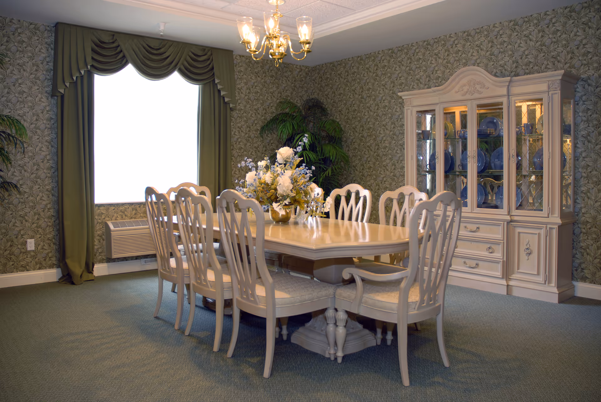 A formal dining room with a rectangular wooden dining table surrounded by eight matching chairs. The table has a floral centerpiece. Behind the table is a large china cabinet displaying blue and white dishware. The room has green patterned wallpaper, green carpet, and a window with green drapes. A chandelier with five lights hangs from the ceiling.