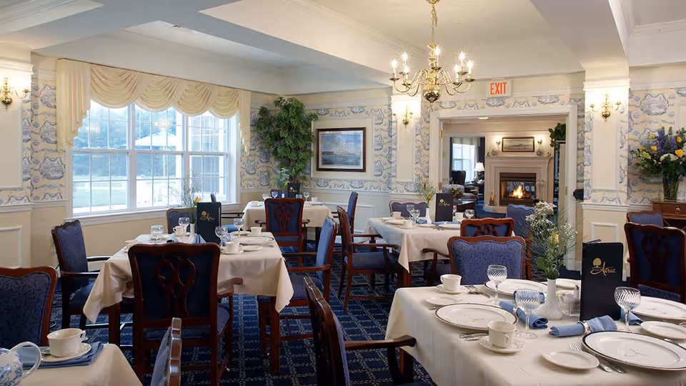 Elegant dining room with tables set with white tablecloths, plates, cups, and glassware. The room features blue upholstered chairs, patterned wallpaper, a large window with draped curtains, a chandelier, and a fireplace visible through an open doorway.