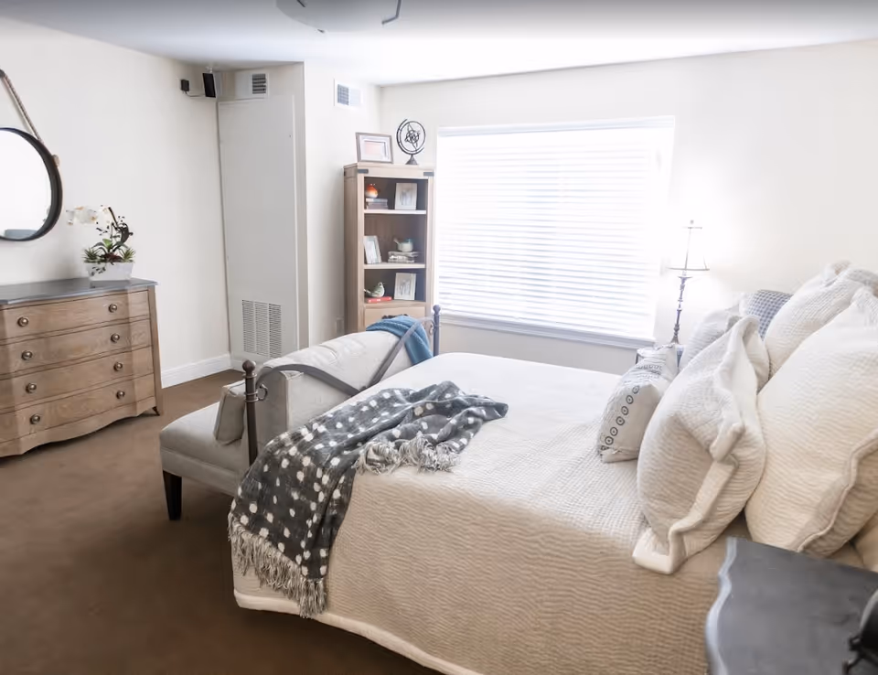 Bright, well-lit bedroom with a made bed topped with multiple pillows, a bench at the foot, a wooden dresser and bookshelf by a large window.