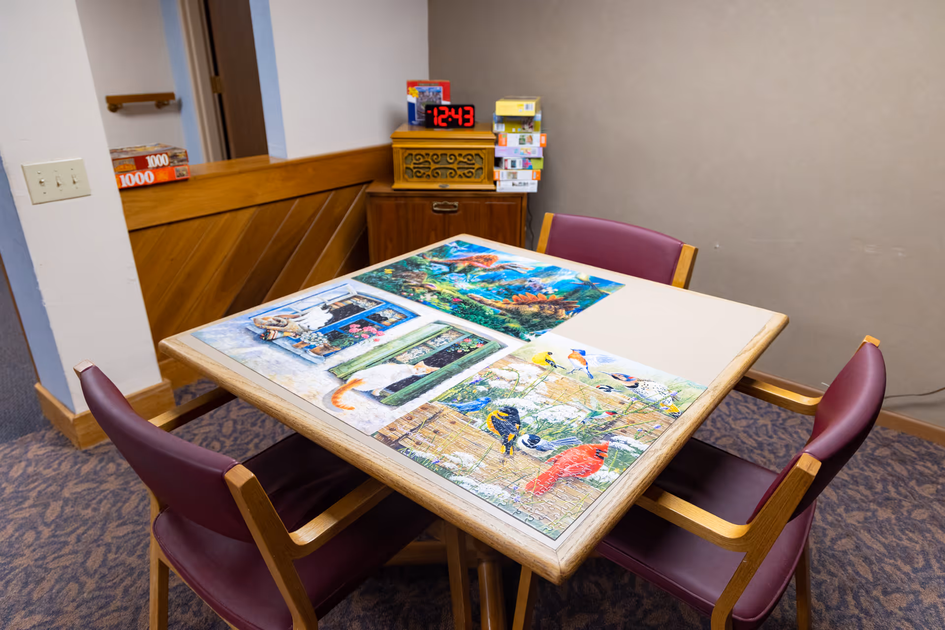 A small table with four maroon chairs around it, featuring three completed colorful jigsaw puzzles depicting birds, a cat by a door, and an underwater scene. In the background, there is a wooden cabinet with a digital clock showing 12:43 and several stacked puzzle boxes.