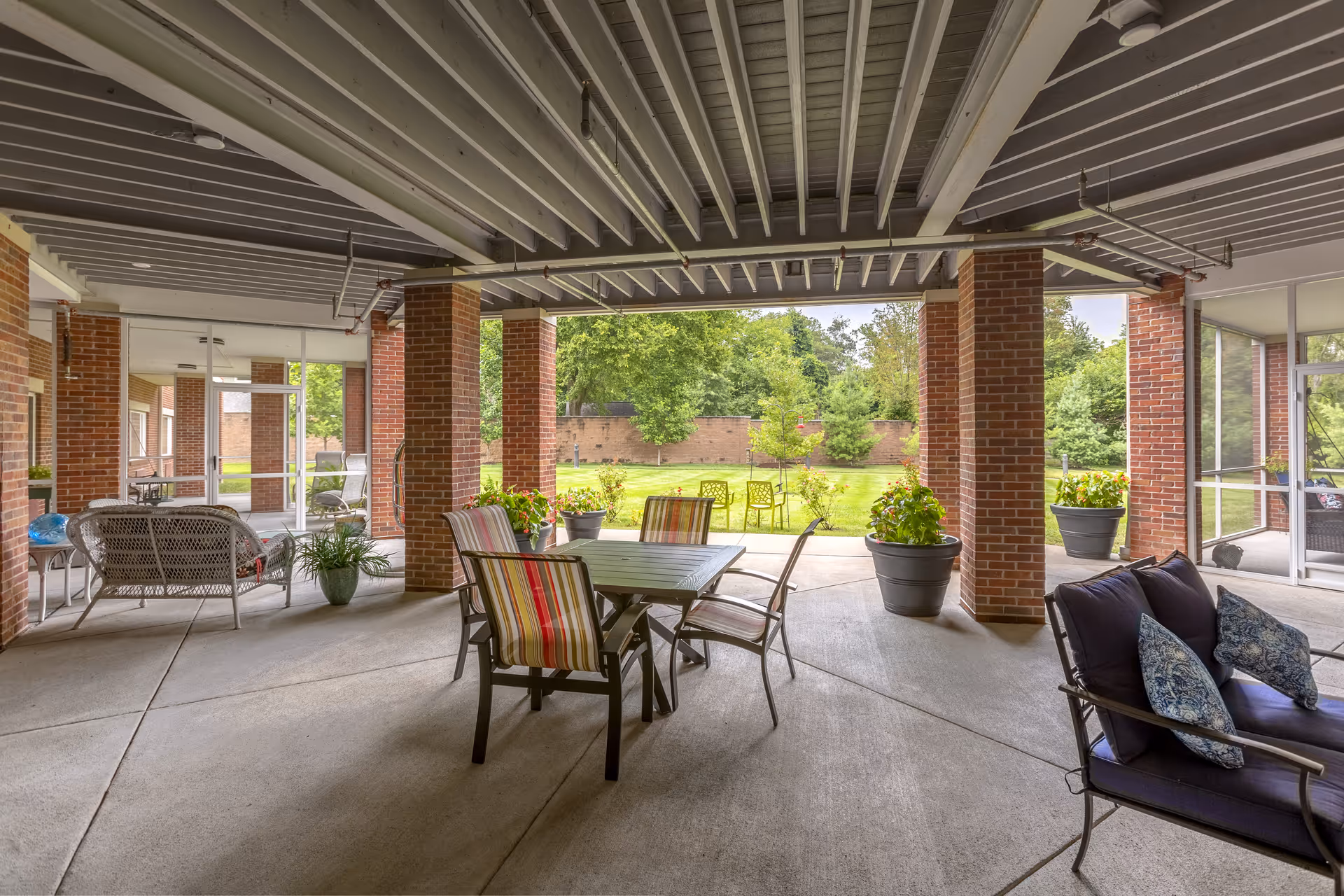 Covered outdoor patio area with brick pillars and a concrete floor. The space is furnished with a table and chairs with striped cushions, a cushioned bench with blue pillows, and several potted plants. Beyond the patio is a grassy area with trees and a brick wall in the background.