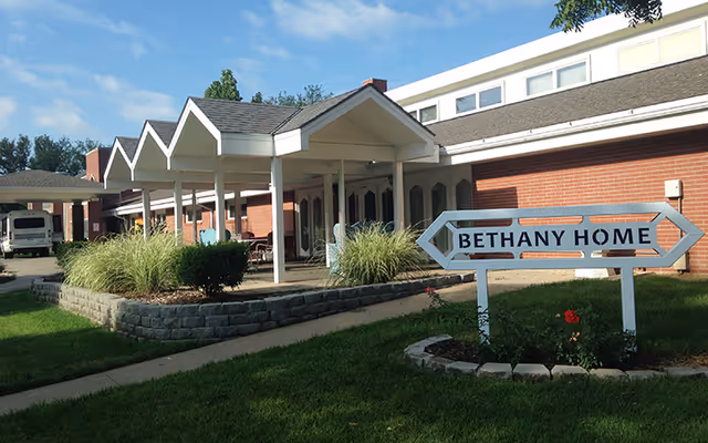 Exterior view of a single-story brick building with a covered entrance supported by white columns. There is a sign in front of the building that reads 'BETHANY HOME' with an arrow pointing to the right. The area around the building has green grass, shrubs, and a small flower bed.