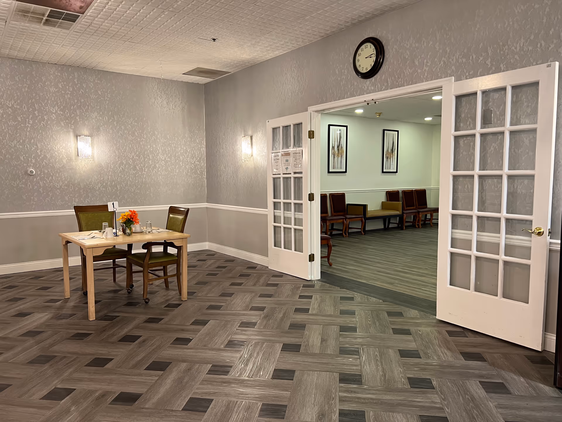 Small dining area with a table set for two and open French doors leading to a seating area with chairs and a wall clock above.
