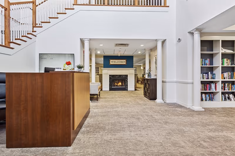 Interior view of a senior living facility lobby with a wooden reception desk on the left, a staircase with wooden railings above, a fireplace with a 'WELCOME' sign above it in the center, and built-in bookshelves filled with books on the right.