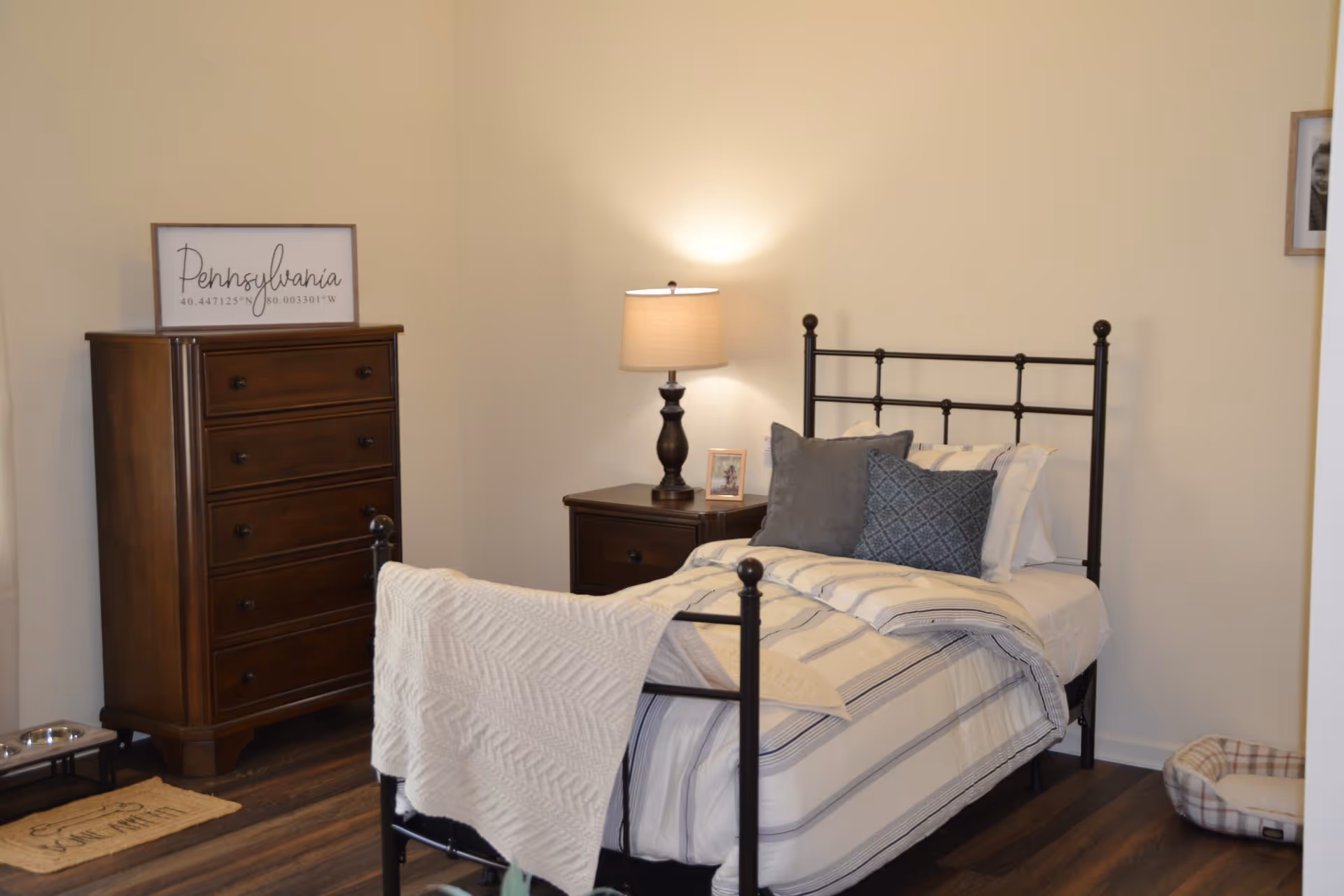 Well-lit single bedroom with a metal-framed bed dressed in striped bedding, a nightstand with a lamp, and a tall wooden dresser.
