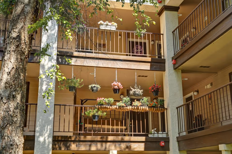 View of an interior courtyard of a multi-level senior living facility with balconies decorated with hanging flower pots and plants. The balconies have brown railings and the walls are beige. A large tree is visible on the left side of the image.