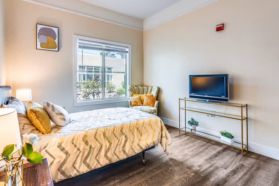 A bright and cozy bedroom with a large window showing outdoor greenery. The room features a bed with patterned bedding and multiple pillows, a wooden nightstand with a lamp and a small plant, a patterned armchair with two orange cushions, and a flat-screen TV on a gold metal stand with two small potted plants underneath. The walls are light-colored and there is a small abstract painting hanging near the window.