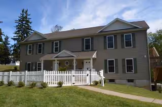 Two-story senior living facility with white picket fence, green lawn, and front entrance.