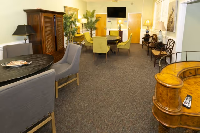 A cozy interior room with several seating areas including a round table with gray chairs in the foreground and a round table with green chairs in the background. The room features wooden furniture, potted plants, lamps, framed artwork on the walls, and a wall-mounted flat screen TV. The carpet is dark patterned and the walls are light-colored.