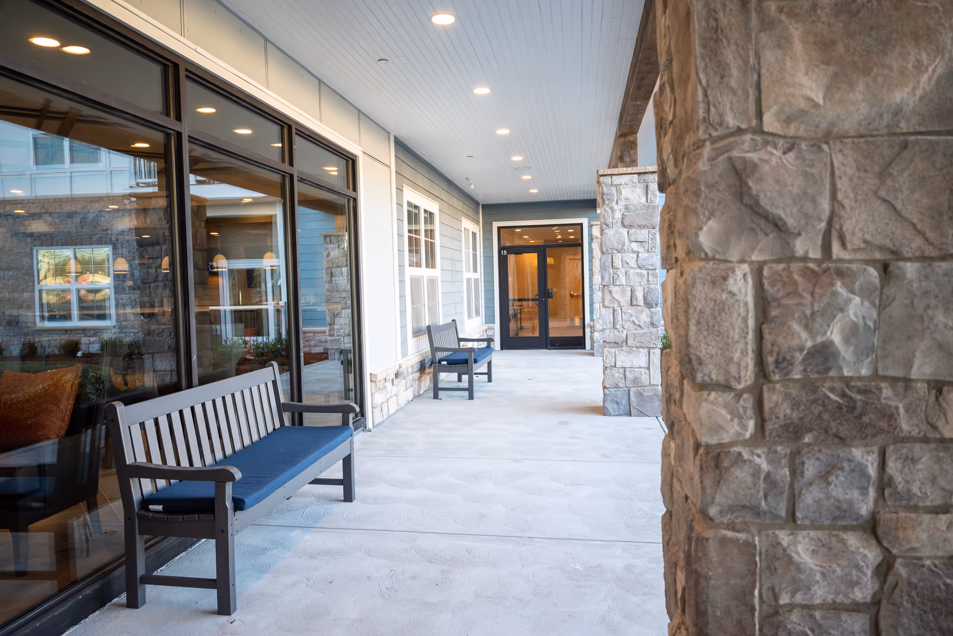 Covered outdoor walkway with stone pillars and benches with blue cushions along the side of a building, large windows reflecting the interior, and a glass door entrance at the end.