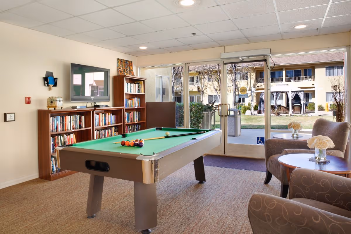 A bright common room with a pool table, bookshelves, chairs, and glass doors leading to a courtyard.