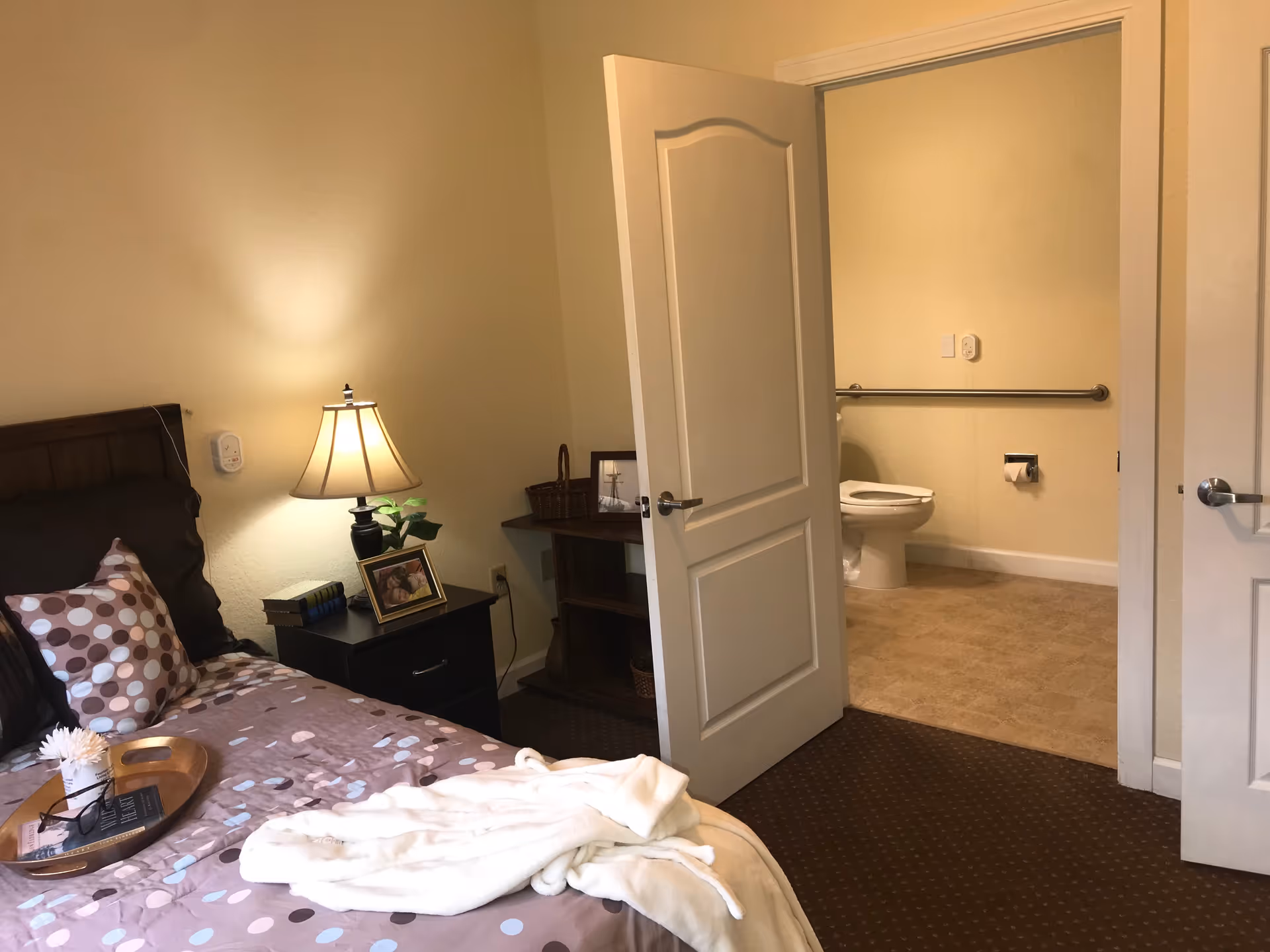 A cozy bedroom with a bed featuring a brown and polka dot patterned comforter and pillows. A nightstand with a lamp, books, a framed photo, and a small plant is next to the bed. The bedroom opens into a bathroom with a visible toilet, grab bar, and toilet paper holder. The walls are painted light yellow and the floor in the bedroom is carpeted while the bathroom floor has tiles.
