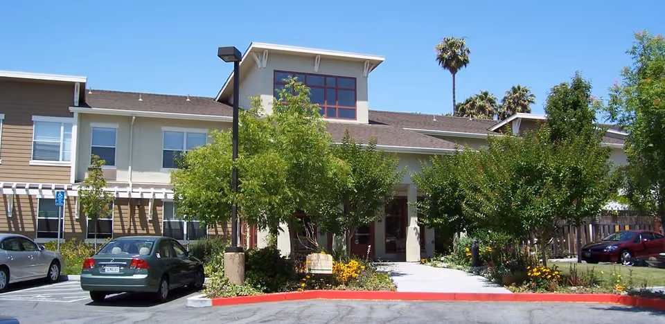 Exterior view of a two-story senior living facility building with a beige and brown facade, surrounded by trees and landscaping. There are cars parked in front, a sidewalk leading to the entrance, and a clear blue sky above.