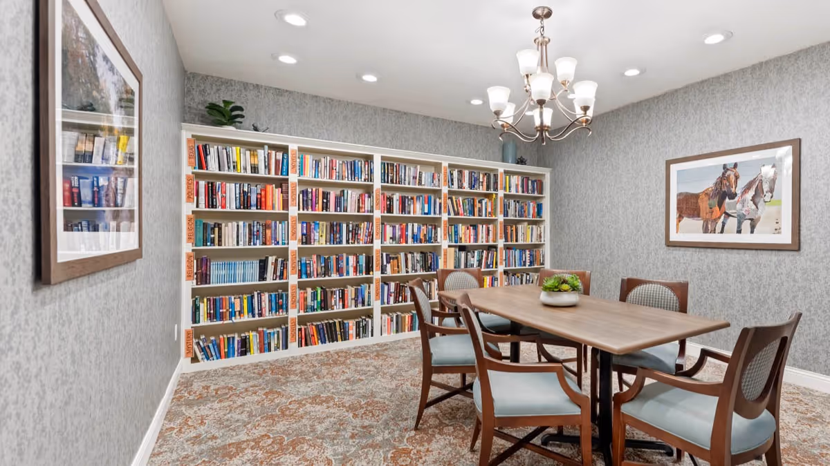A cozy library room with a large bookshelf filled with books along the back wall. In front of the bookshelf is a wooden table surrounded by six cushioned chairs. The room has patterned carpet, gray textured walls, a chandelier hanging from the ceiling, and framed artwork on the walls.