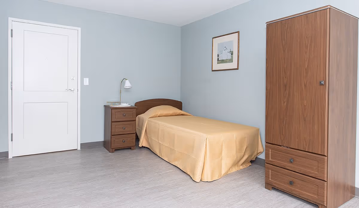 A simple bedroom with a single bed covered in a beige bedspread, a wooden nightstand with a lamp and a few items on it, a wooden wardrobe, and a framed picture hanging on a light blue wall.