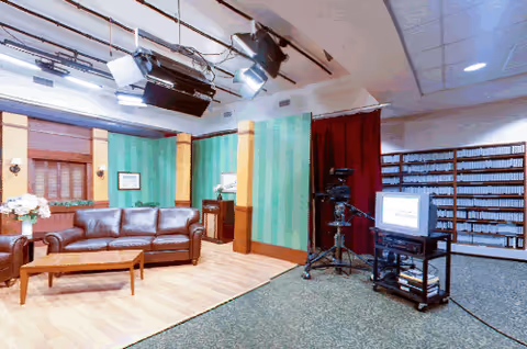 A studio set designed to look like a living room with a brown leather couch, wooden coffee table, green striped wallpaper, and framed pictures on the wall. Professional studio lighting and a video camera on a tripod are positioned in front of the set. To the right, there is a TV on a stand and shelves filled with VHS tapes or similar media.