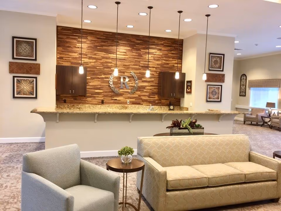 A cozy seating area in a memory care facility featuring a beige patterned sofa, a light gray armchair, and a small round wooden table with a small plant. Behind the seating area is a counter with a granite countertop and a wood-paneled wall with a metallic letter 'R' and laurel wreath decoration. Pendant lights hang from the ceiling, and framed artwork decorates the walls.