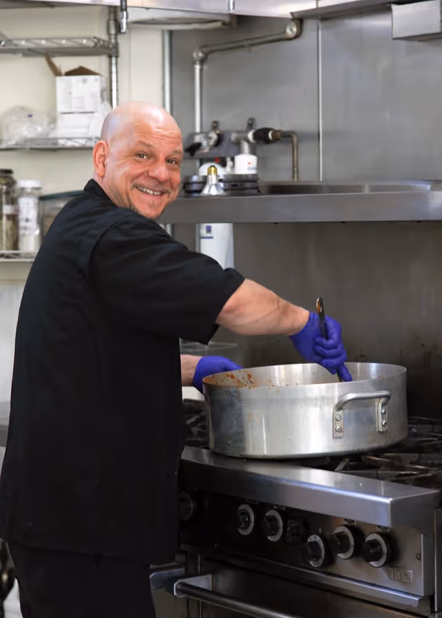 A smiling chef wearing a black uniform and purple gloves stirring food in a large metal pot on a stove in a commercial kitchen.