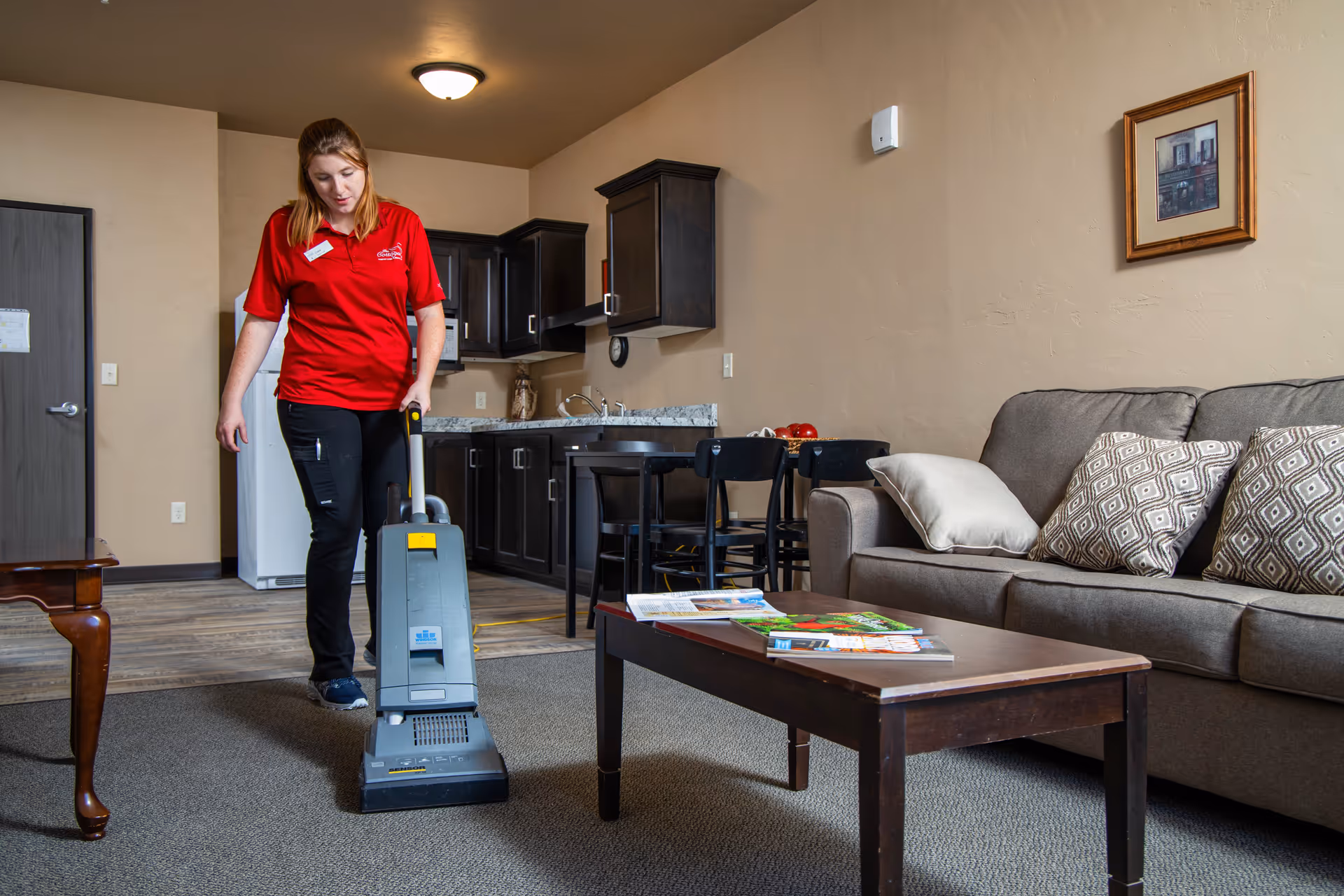 A staff member in a red shirt vacuums the carpet in a furnished living room with a sofa, coffee table, and kitchenette.