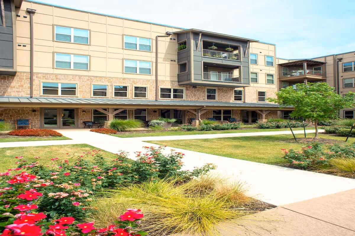 Exterior view of a multi-story senior living facility building with balconies, surrounded by landscaped gardens featuring green grass, flowering plants, and small trees under a clear sky.