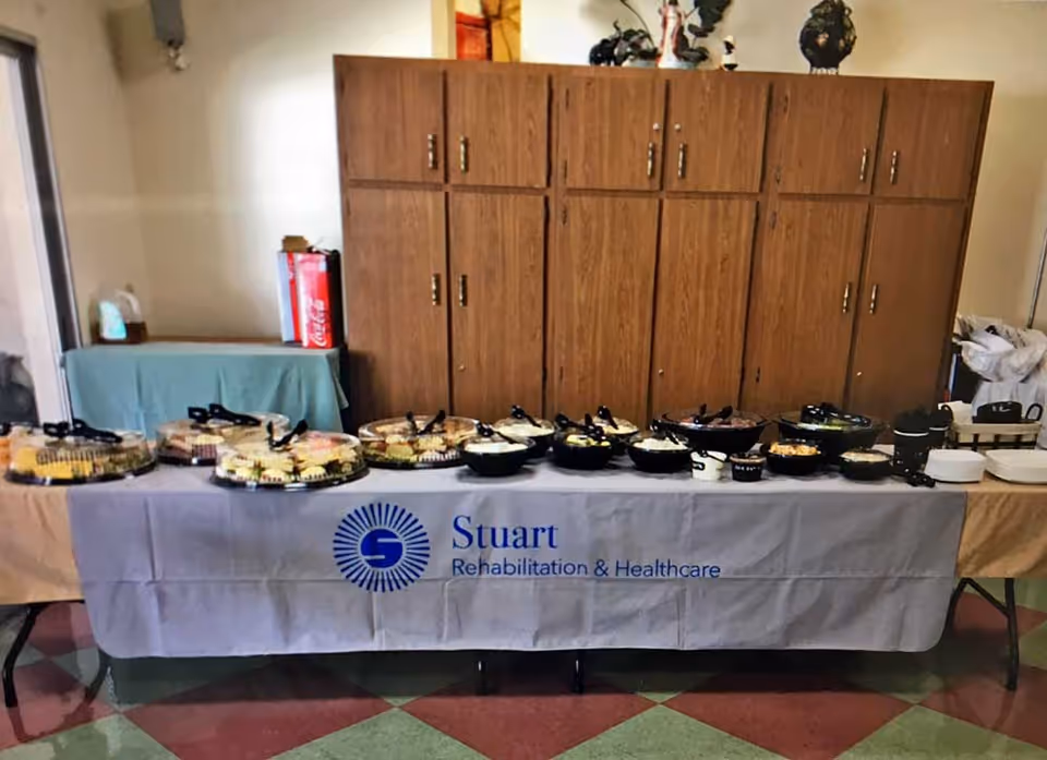 Buffet table with assorted covered platters and bowls set in front of wooden cabinets, draped with a tablecloth reading 'Stuart Rehabilitation & Healthcare'.