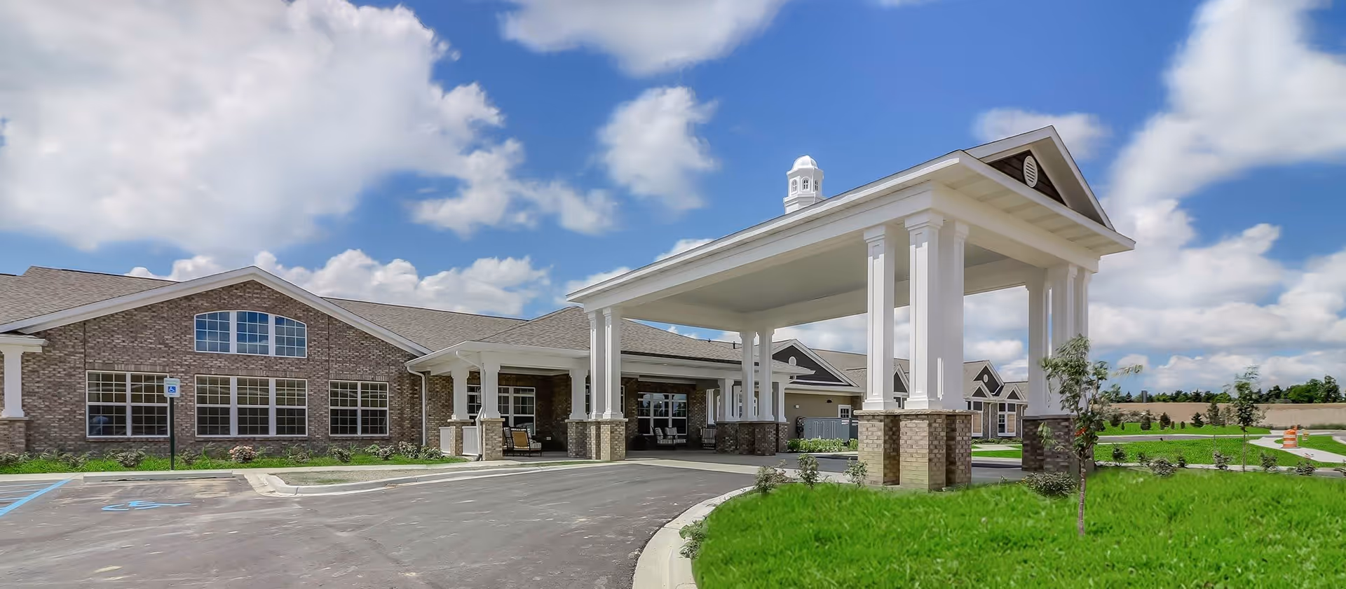 Exterior view of Springvale Assisted Living & Memory Care facility showing a large covered entrance with white columns and brick base, a paved driveway, green lawn, and a partly cloudy blue sky.