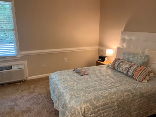 A bedroom with a bed covered in a light blue patterned bedspread and several pillows, including a long cylindrical one. There is a small wooden nightstand with a lamp and some items on it. A window with blinds and an air conditioning unit below it is visible on the left side. The walls are painted beige with white trim.