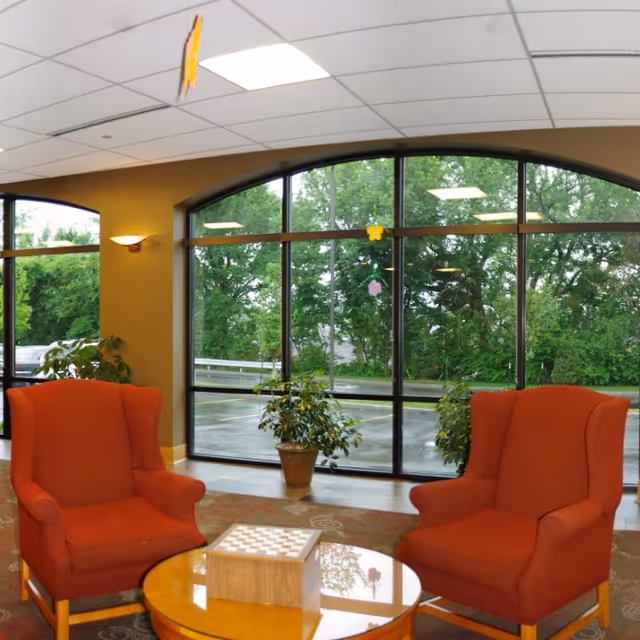 Lobby seating area with two red armchairs around a round table with a checkerboard in front of large arched windows looking out to trees.