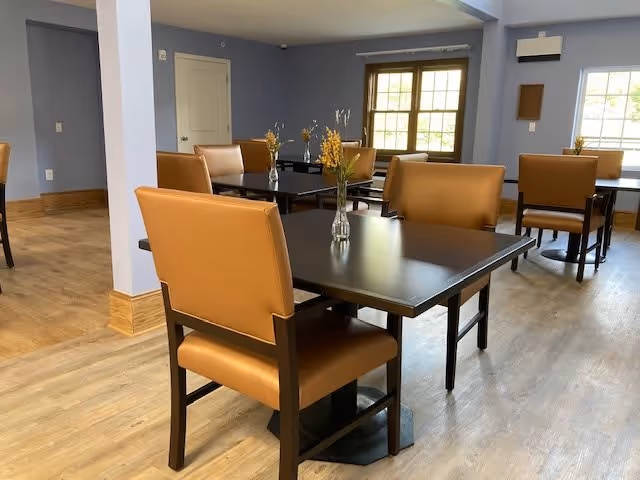 A dining area with several dark wooden tables and tan cushioned chairs. Each table has a small vase with yellow flowers. The room has light blue walls, a window letting in natural light, and wood flooring.