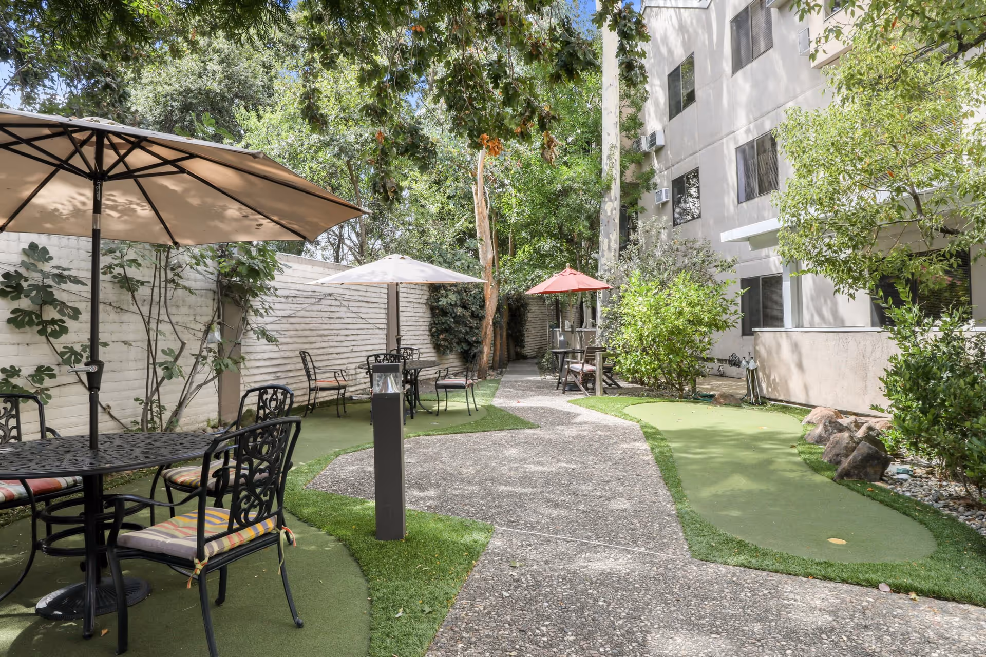 Outdoor patio area at The Heritage Downtown with several black metal tables and chairs, each shaded by large umbrellas. The area is surrounded by greenery including trees and bushes, with a concrete pathway running through the middle. The building exterior is visible on the right side.