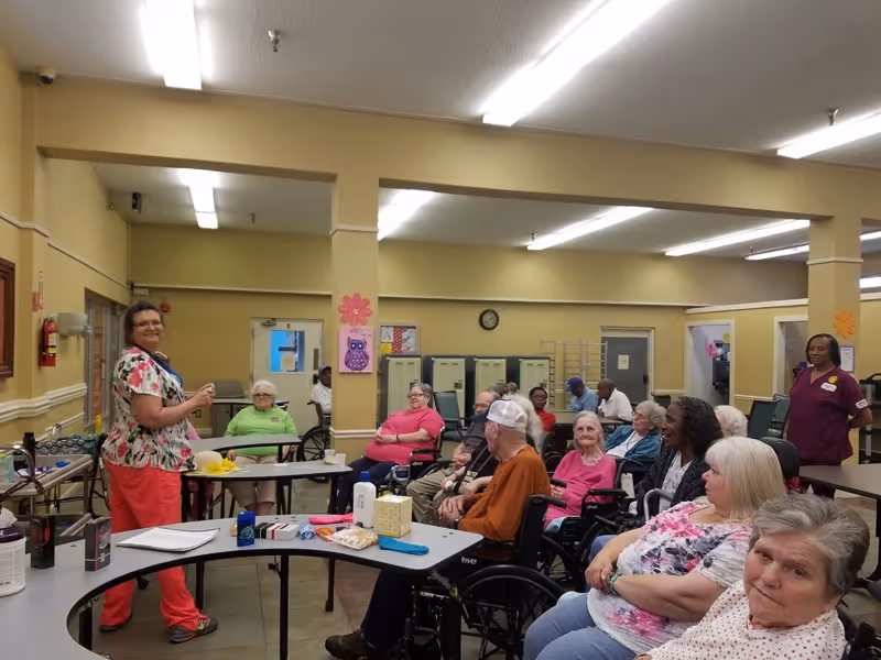 A group of elderly residents, some in wheelchairs, seated around tables in a well-lit room with beige walls. Two staff members stand nearby, one in a floral top and red pants speaking to the group, and another in a maroon uniform standing at the back. The room has lockers, a clock on the wall, and colorful decorations including a pink flower and an owl drawing.