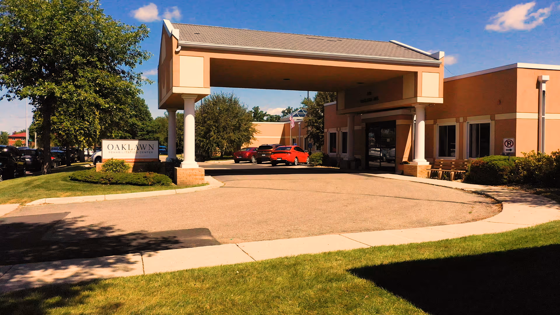 Front entrance of Oaklawn Rehabilitation Center with a covered porte-cochere, driveway, cars, and landscaping on a sunny day.