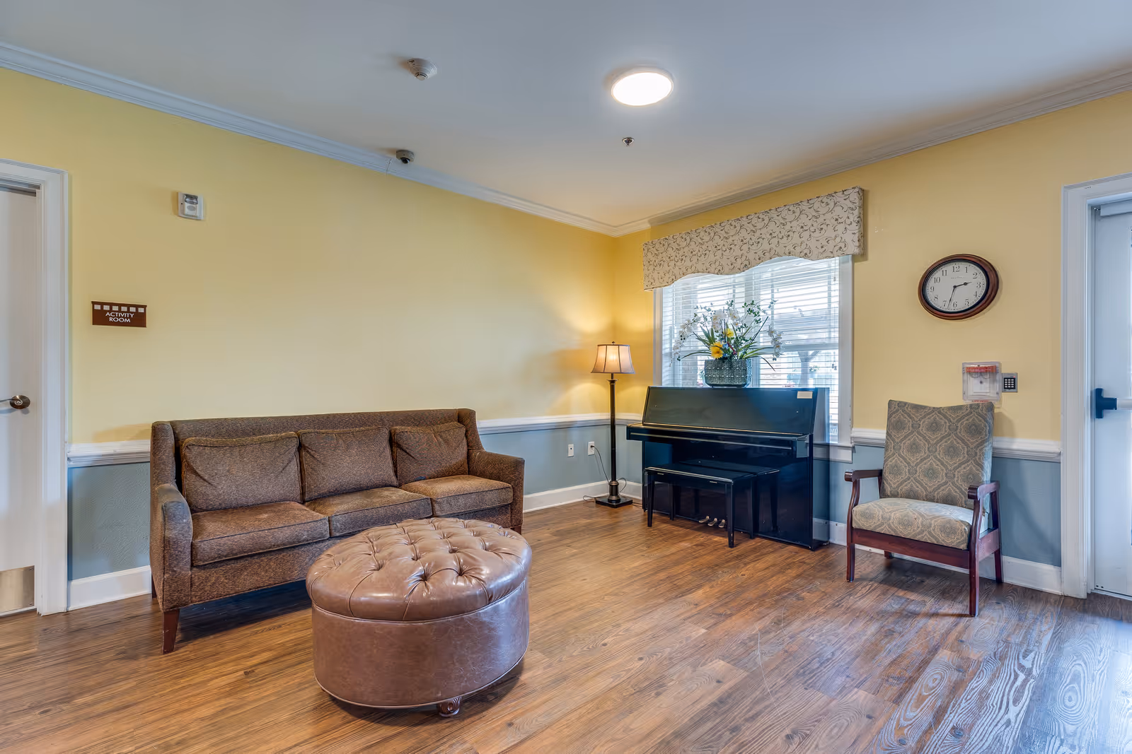 A cozy room with a brown sofa, a round tufted leather ottoman, a patterned armchair, a black upright piano with a floral arrangement on top, a standing lamp, and a wall clock. The walls are painted yellow and blue with white trim, and there is a window with blinds and a decorative valance.