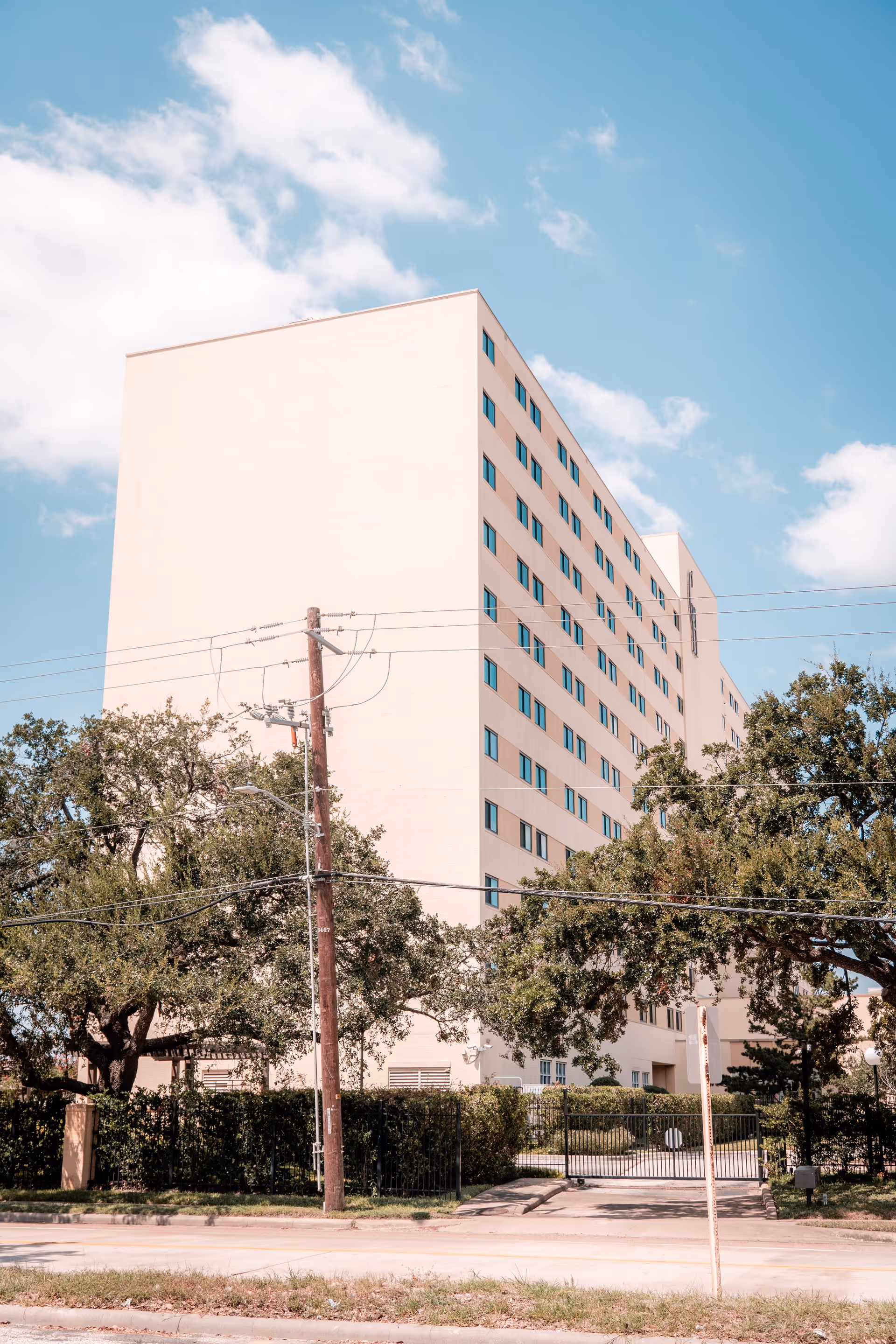 A tall, light-colored multi-story building with many windows, surrounded by trees and a gated entrance, under a partly cloudy blue sky.